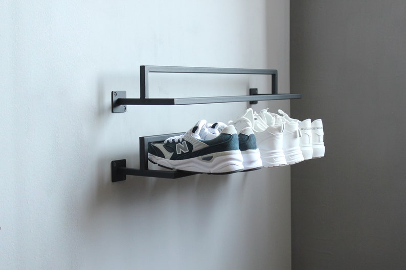 May include: A black metal shoe rack with two shelves mounted on a white wall. The top shelf has four pairs of white sneakers, and the bottom shelf has one pair of green and white sneakers.