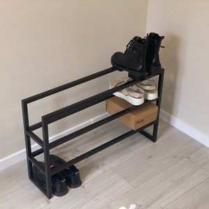 May include: Black metal shoe rack with three tiers. The top tier has two pairs of black boots and a pair of white sandals. The middle tier has a brown cardboard box. The bottom tier has two pairs of black shoes.
