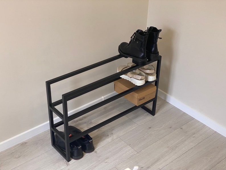 May include: Black metal shoe rack with three tiers. The top tier has two pairs of black boots and a pair of white sandals. The middle tier has a brown cardboard box. The bottom tier has two pairs of black shoes.