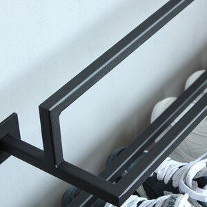 May include: Black metal shoe rack with two shelves mounted to a wall. The rack is holding two pairs of sneakers, one pair is white with black accents and the other pair is blue and white.