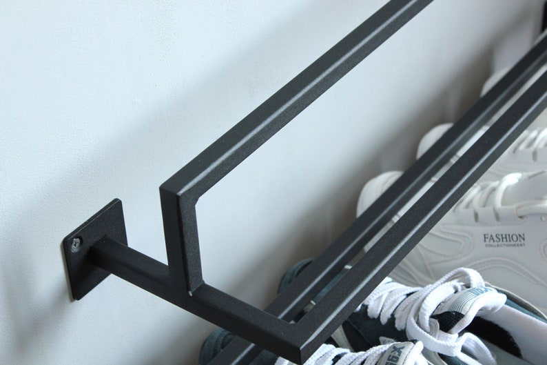 May include: Black metal shoe rack with two shelves mounted to a wall. The rack is holding two pairs of sneakers, one pair is white with black accents and the other pair is blue and white.