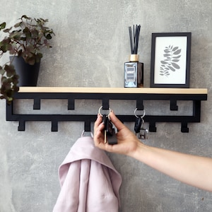 Puede incluir: Una estantería de pared de metal negro con ganchos, una repisa superior de madera y una pequeña planta en maceta. Las llaves cuelgan de los ganchos. Un marco de imagen con un diseño de hoja está en la estantería.