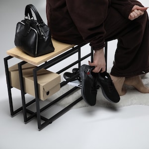 以下が含まれることがあります： 黒い革のハンドバッグとシルバーのジッパーが、黒い金属フレームの木製ベンチの上に置かれています。 下には2つの段ボールの靴箱があります。 赤と白のアクセントが付いた黒いスニーカーを人が持っています。