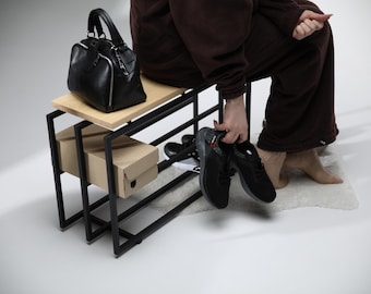 May include: A black leather handbag with silver zips rests on a wooden bench with a black metal frame. Two cardboard shoe boxes are below. A person holds a pair of black trainers with red and white accents.