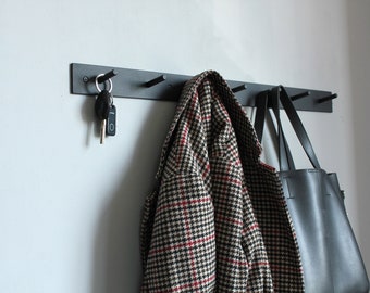 Könnte beinhalten: Ein schwarzer, wandmontierter Kleiderhaken aus Metall mit fünf Haken. Eine karierte Jacke hängt an einem Haken, eine schwarze Ledertasche hängt an einem anderen und ein Schlüsselbund hängt an einem dritten Haken.