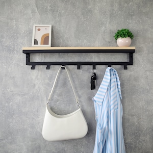 Puede incluir: Un estante de pared con una repisa de madera clara sobre una estructura de metal negro con siete ganchos. Un bolso blanco, un juego de llaves y una camisa a rayas azul claro y blanco cuelgan de los ganchos. Un marco pequeño y una planta están encima.