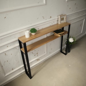 May include: A narrow console table featuring two rectangular light wood shelves supported by a black metal frame. The top shelf holds a small white cylindrical container, a green potted plant, and a small picture frame. Two books rest on the lower shelf.