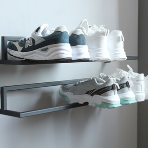 May include: A black metal shoe rack with two shelves mounted on a wall. The top shelf holds two pairs of sneakers, one pair is white and the other is blue and white. The bottom shelf holds three pairs of white sneakers.
