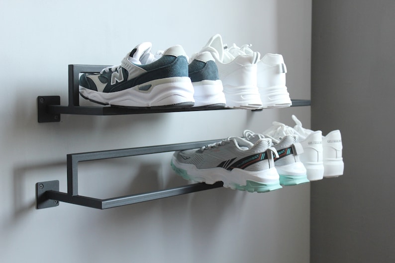 May include: A black metal shoe rack with two shelves mounted on a wall. The top shelf holds two pairs of sneakers, one pair is white and the other is blue and white. The bottom shelf holds three pairs of white sneakers.