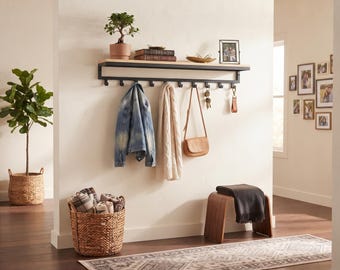 Puede incluir: Un organizador de entrada montado en la pared con un estante de madera clara sobre un marco de metal negro con ocho ganchos. El estante sostiene una pequeña planta en maceta, libros y un marco de fotos. Debajo, una chaqueta, bufanda y bolso.