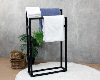 May include: Black metal towel rack with three bars for hanging towels. Two white towels and one blue towel are hanging on the rack. The rack is standing on a white rug in front of a gray wall.