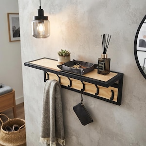 Puede incluir: Un estante de pared para recibidor con marco de metal negro y superficie de madera clara. La unidad incluye cinco ganchos de metal negro debajo para colgar objetos. Sobre el estante hay una bandeja de tela gris, una suculenta y un difusor.