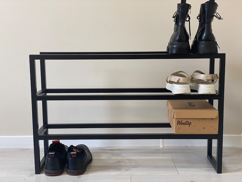 May include: Black metal shoe rack with three shelves. The top shelf has two pairs of black boots. The middle shelf has two pairs of sandals. The bottom shelf has a box labeled "Weestep" and two pairs of black shoes.
