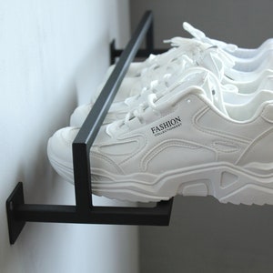 May include: A black metal wall-mounted shoe rack with three pairs of white sneakers. The sneakers have black laces and the text "FASHION COLLECTIONEVERY" is printed on the side of one of the sneakers.