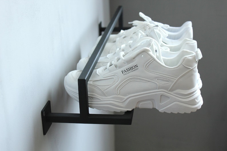 May include: A black metal wall-mounted shoe rack with three pairs of white sneakers. The sneakers have black laces and the text "FASHION COLLECTIONEVERY" is printed on the side of one of the sneakers.