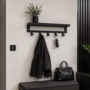 Puede incluir: Un perchero de pared negro con un estante superior plano y cinco ganchos. El perchero sostiene una chaqueta vaquera oscura y un juego de llaves. El estante sostiene una pequeña planta en maceta dentro de un recipiente negro con estrías.
