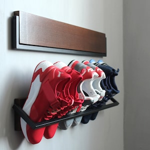 May include: A black metal shoe rack with six pairs of shoes, including red, white, and blue sneakers, mounted on a white wall. The rack is attached to a wooden shelf.