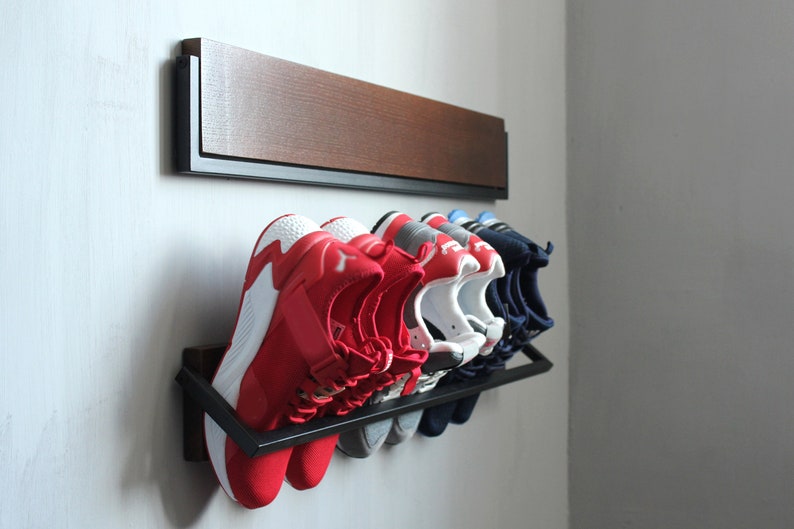 May include: A black metal shoe rack with six pairs of shoes, including red, white, and blue sneakers, mounted on a white wall. The rack is attached to a wooden shelf.