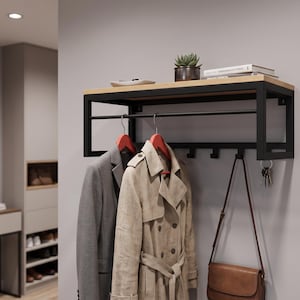 Peut inclure: Un porte-manteau mural composé d'un cadre en métal noir et d'une étagère supérieure en bois clair. L'unité comprend une tringle horizontale avec des cintres rouges portant un blazer gris et un trench-coat beige, ainsi que des crochets noirs.