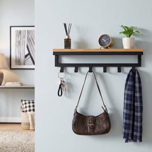 Puede incluir: Un estante de pared con una superficie superior de madera natural y un marco de metal negro mate. La unidad incluye seis ganchos integrados en la parte inferior para colgar objetos, que actualmente sostienen un llavero, un bolso de cuero marrón y una bufanda.
