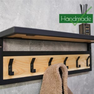 Puede incluir: Estante de pared de metal negro y madera clara con seis ganchos. El marco es negro y la parte trasera de madera natural. Una toalla marrón cuelga de uno de los ganchos. La palabra "Handmade" está en un rectángulo verde.