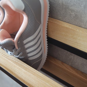 May include: A pair of gray sneakers with white stripes and pink accents are placed on a wooden shelf with black metal supports.