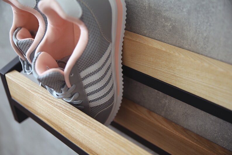 May include: A pair of gray sneakers with white stripes and pink accents are placed on a wooden shelf with black metal supports.