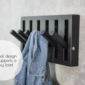 May include: A black wooden coat rack with six angled hooks, holding a denim jacket. The rack has a rectangular design with vertical slots. Text on the image reads "Original design and supports a heavy load."