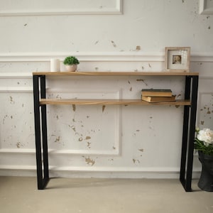 Puede incluir: Una mesa consola de metal negro con dos estantes de madera. La mesa tiene un diseño sencillo y es perfecta para un pasillo o una entrada. La mesa está decorada con una pequeña planta en maceta, una foto enmarcada y una pila de libros.