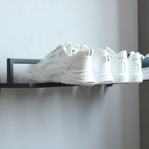 May include: A black metal shoe rack mounted on a wall with six pairs of shoes. Five pairs are white sneakers and one pair is blue and white sneakers.