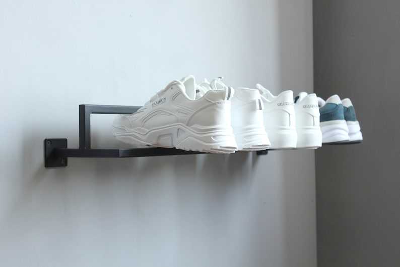May include: A black metal shoe rack mounted on a wall with six pairs of shoes. Five pairs are white sneakers and one pair is blue and white sneakers.