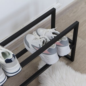 May include: A black metal shoe rack with two tiers holds four pairs of sneakers. The sneakers are white with grey and pink accents. The rack is on a wooden floor with a white fluffy rug.