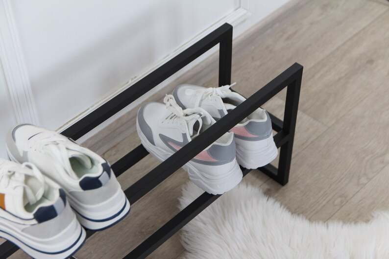 May include: A black metal shoe rack with two tiers holds four pairs of sneakers. The sneakers are white with grey and pink accents. The rack is on a wooden floor with a white fluffy rug.