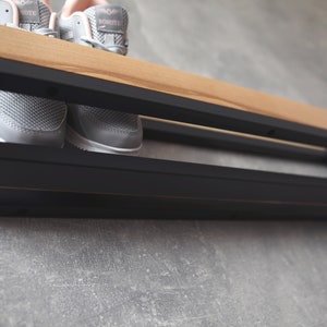 May include: A black metal shoe rack with a wooden shelf, mounted on a gray wall. Two pairs of gray sneakers are on the shelf.