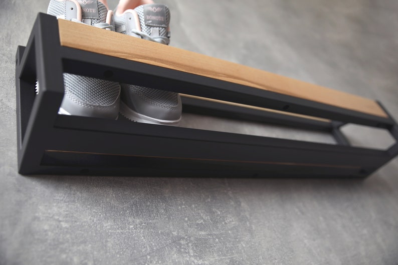 May include: A black metal shoe rack with a wooden shelf, mounted on a gray wall. Two pairs of gray sneakers are on the shelf.