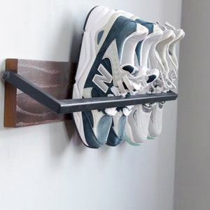 May include: A wall-mounted shoe rack with a dark wood base and a black metal bar. Four pairs of sneakers are hanging on the bar, including a pair of blue and white sneakers.