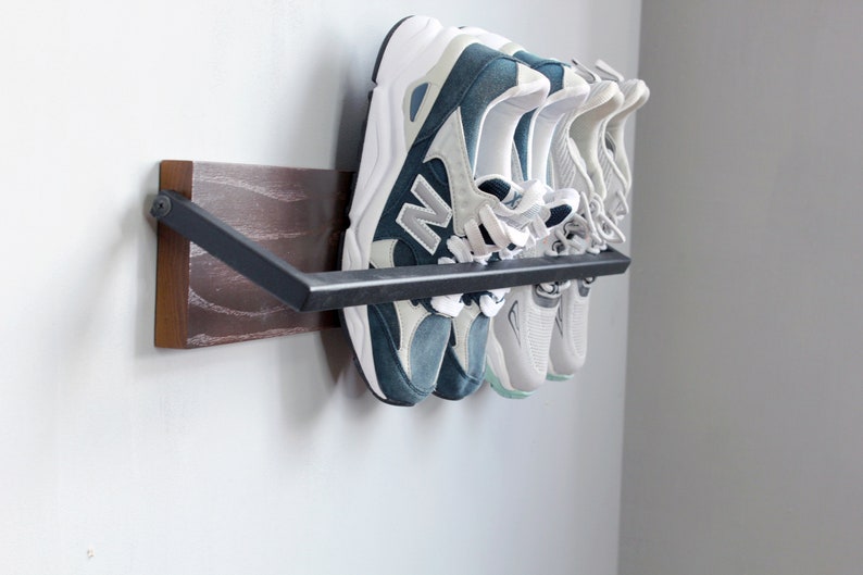 May include: A wall-mounted shoe rack with a dark wood base and a black metal bar. Four pairs of sneakers are hanging on the bar, including a pair of blue and white sneakers.