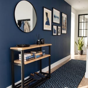 Puede incluir: Una mesa consola rectangular con una superficie de madera clara y un marco de metal negro, que cuenta con dos estantes. El estante superior sostiene una bandeja negra y dos objetos decorativos negros, mientras que el estante medio tiene libros.