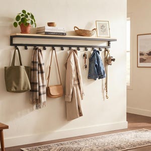 Puede incluir: Un organizador de pared para entrada con un estante de madera clara sostenido por un marco de metal negro con diez ganchos integrados. El estante sostiene una planta en maceta, libros, un cuenco pequeño, una cesta tejida y un cuadro enmarcado.