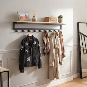 Puede incluir: Un perchero de pared con estante de madera clara sobre una estructura de metal negro con ocho ganchos. Sostiene una chaqueta vaquera negra con parches, un pañuelo estampado y una gabardina beige para un almacenamiento organizado.
