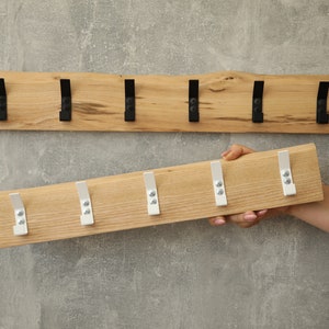 Puede incluir: Dos percheros de madera con ganchos metálicos. Uno está montado en una pared gris, con ganchos negros. El otro, sostenido por una mano, tiene ganchos blancos. Ambos son de madera clara.