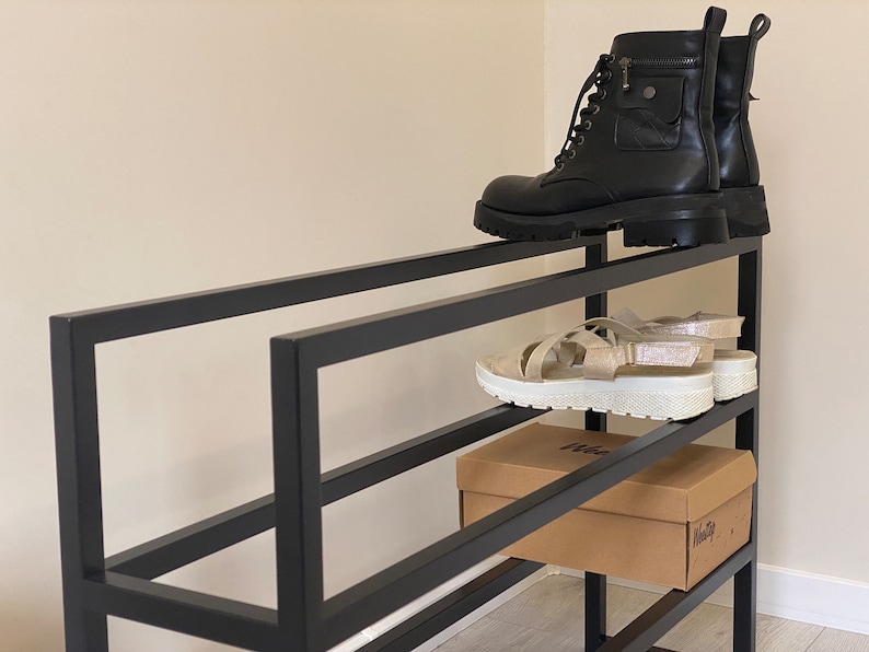 May include: A black metal shoe rack with three shelves. The top shelf has a pair of black boots. The middle shelf has a pair of white sandals and a brown cardboard box with the word "Hello" printed on it. The bottom shelf is empty.