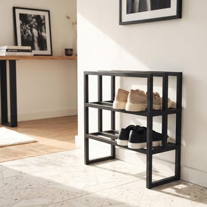 May include: A black metal shoe rack with a rectangular frame design featuring two tiers. The top shelf holds a pair of beige sneakers, while the bottom shelf holds a pair of black sneakers with white soles, positioned against a white wall.