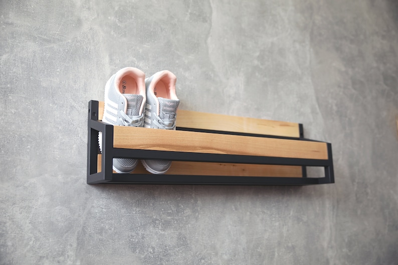 May include: A black metal shoe rack with a wooden shelf, mounted on a gray wall. Two gray sneakers with pink accents are on the shelf.