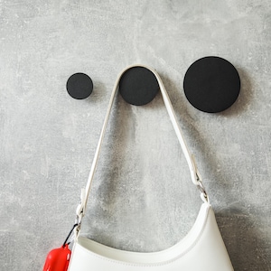 May include: Three black circular wall hooks of varying sizes are mounted on a textured grey wall. A white shoulder bag hangs from the central hook, with a red item attached to the strap. The hooks offer a minimalist storage solution.