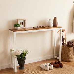 May include: A rectangular console table with a natural wood top and a white metal frame. The table surface holds a small potted plant, a wooden picture frame, a gold tray with sunglasses, and two vases. A black face-shaped planter sits below.