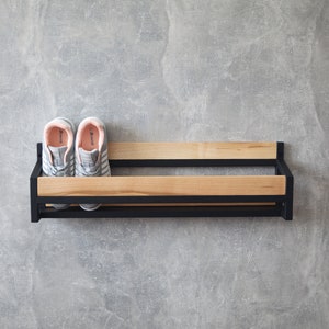 May include: A black metal shoe rack with two wooden shelves. The rack is mounted on a gray wall and has two pairs of gray sneakers with pink accents on the shelves.