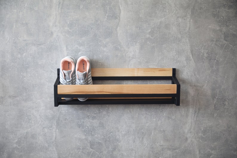 May include: A black metal shoe rack with two wooden shelves. The rack is mounted on a gray wall and has two pairs of gray sneakers with pink accents on the shelves.