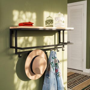 Könnte beinhalten: Ein Wandregal mit einem hellen Holzbrett auf einem schwarzen Metallrahmen. Das Gestell verfügt über eine horizontale Stange und fünf Haken, an denen ein beigefarbener Fedora-Hut und eine hellblaue Jeansjacke mit Blumenstickerei hängen.
