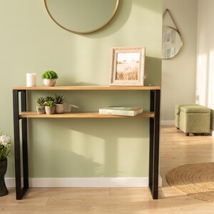 May include: A console table featuring two horizontal light wood shelves supported by a black metal frame. The top shelf holds a white candle, a small green plant, and a framed picture. The lower shelf holds three small potted plants and a book.